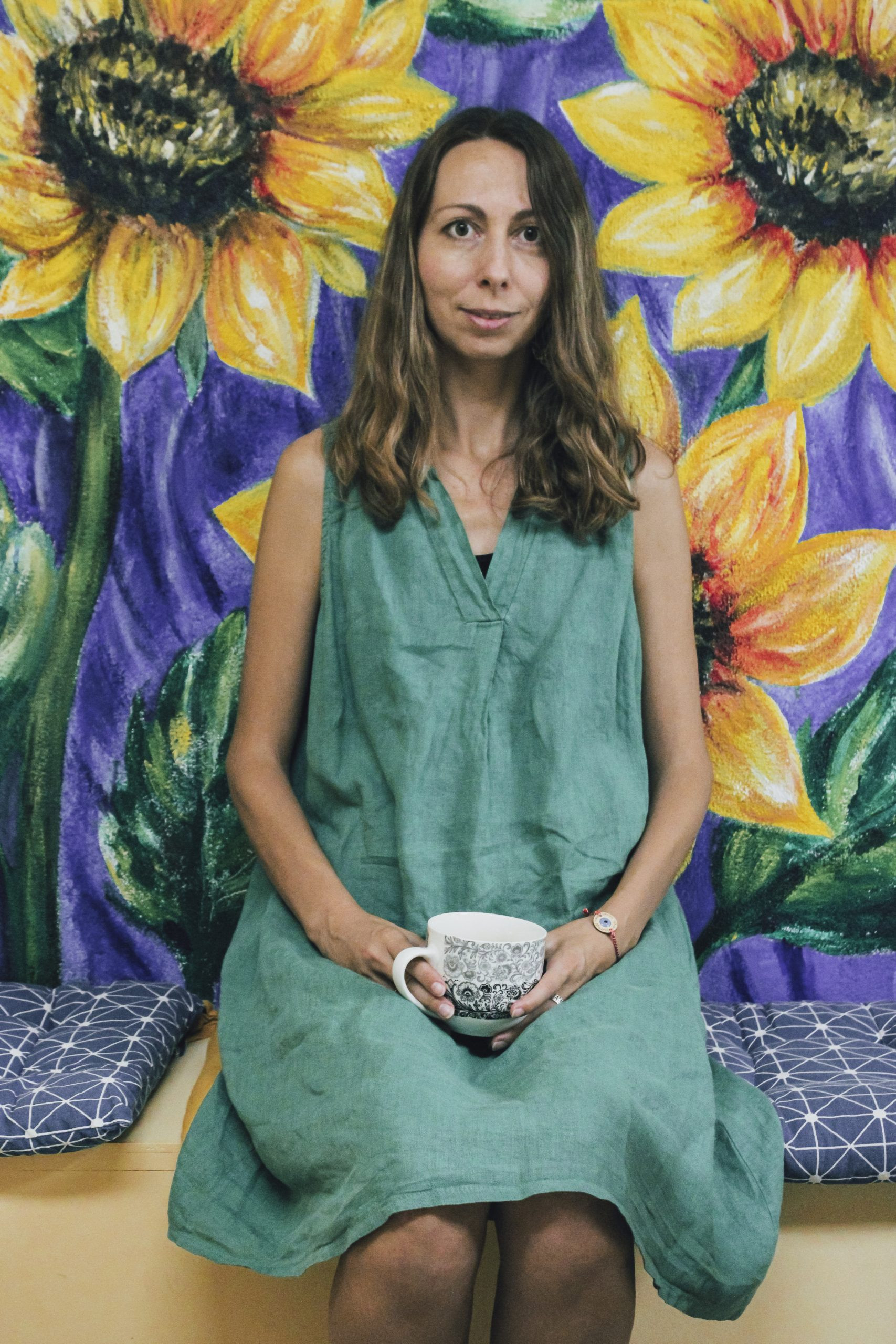 A woman sitting in front of a wall with painted flowers holding a mug.