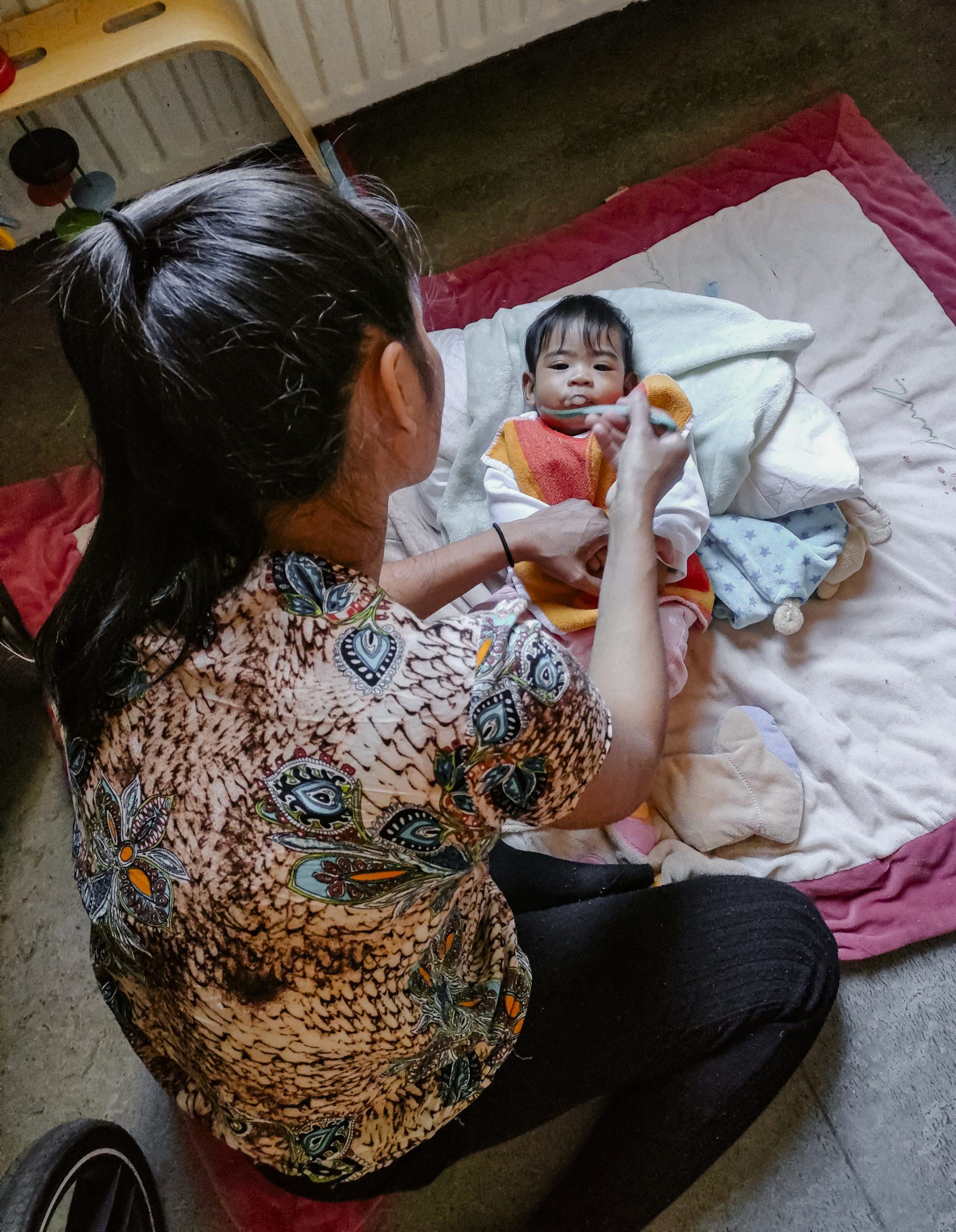 A mother, sitting on the floor, feeding her baby with a spoon.