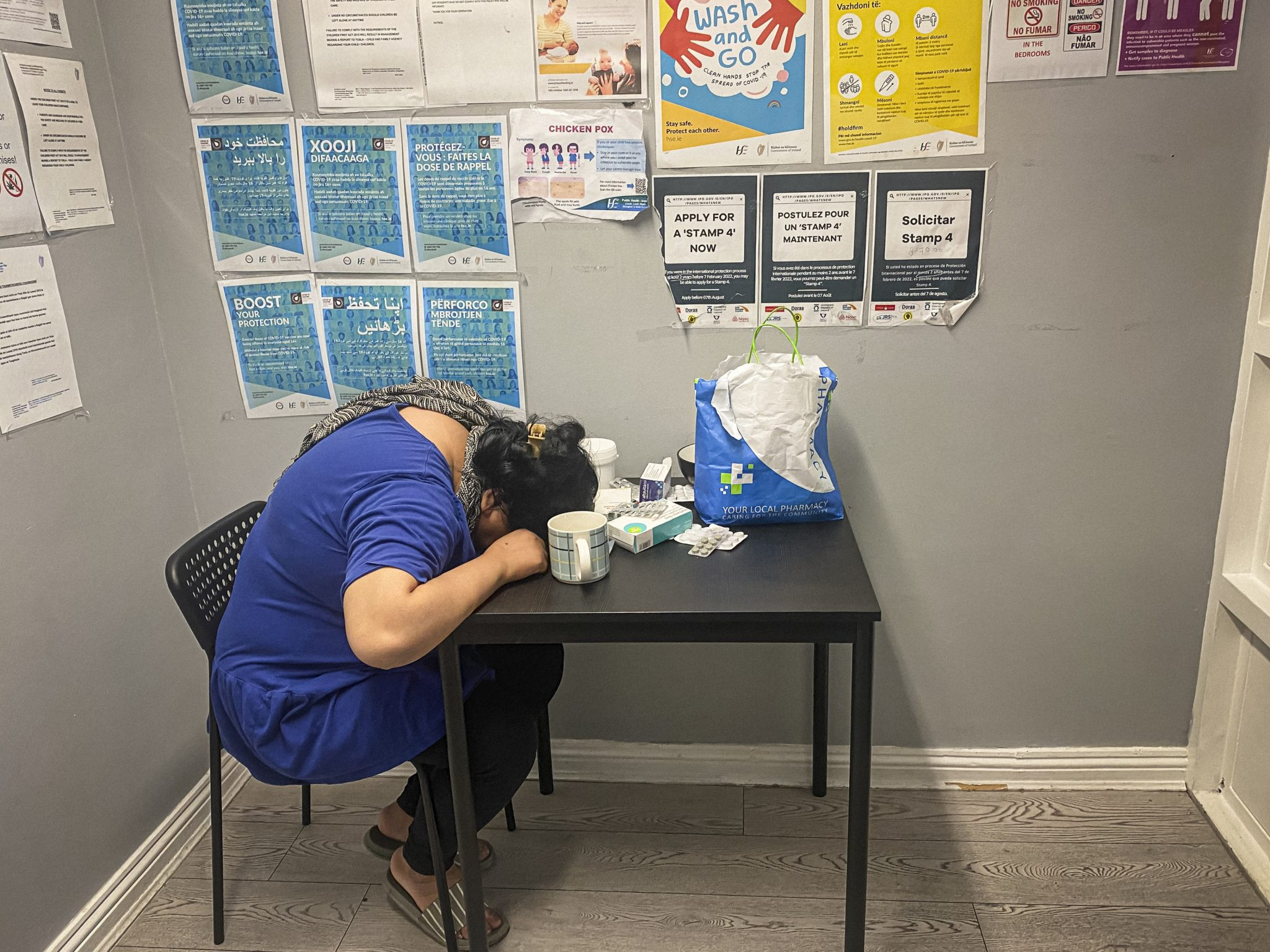 An exhausted and sick woman, sitting on a chair with her head on the table, surrounded by different kinds of medicine.