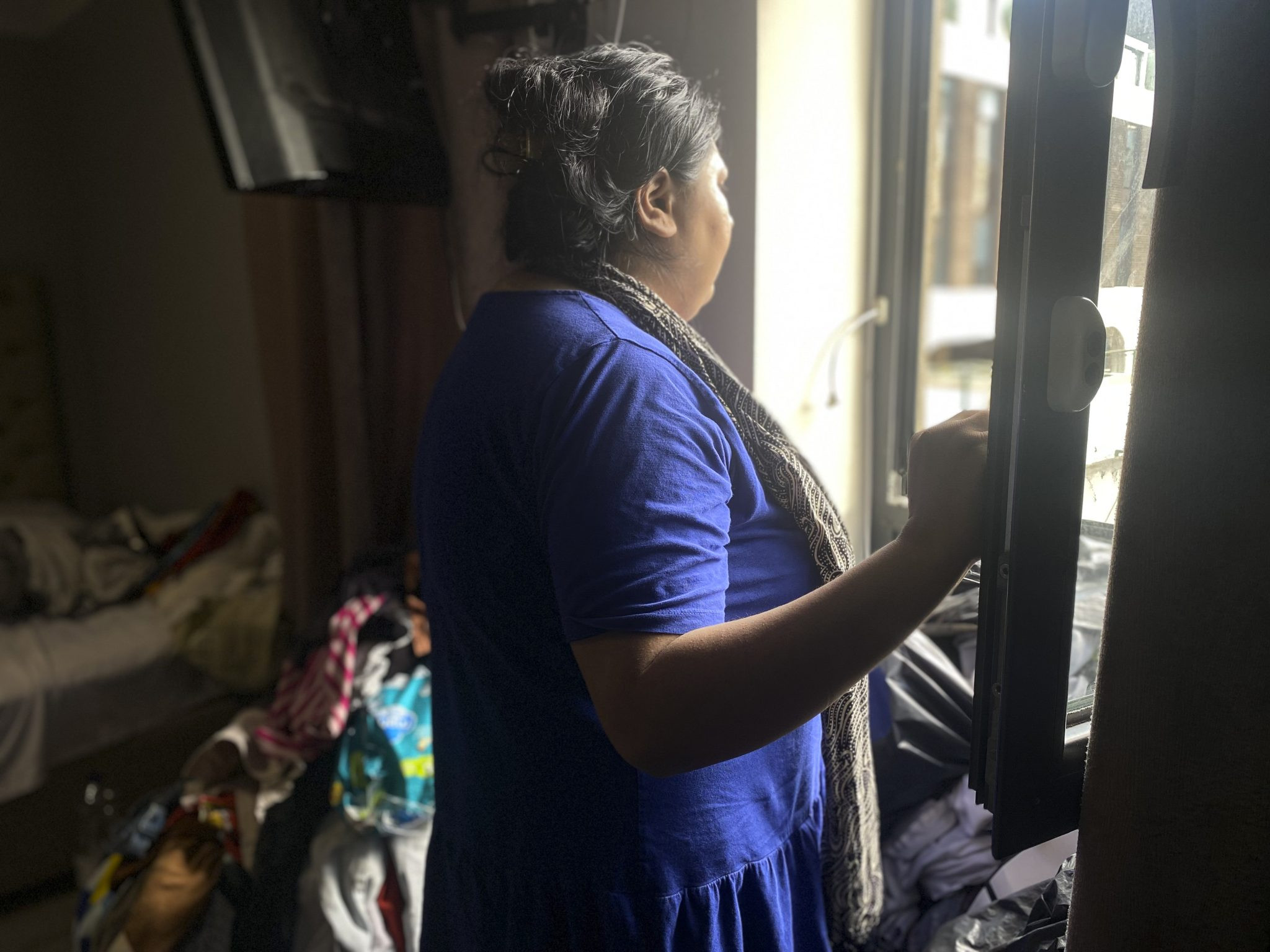 A woman staring out an open window. There is also lots of clutter in the house around her.